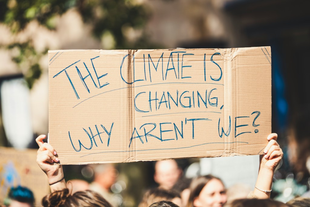 Protesters holding a cardboard sign reading "The Climate Is Changing, Why Aren't We?" during a climate demonstration.