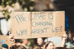 Protesters holding a cardboard sign reading "The Climate Is Changing, Why Aren't We?" during a climate demonstration.
